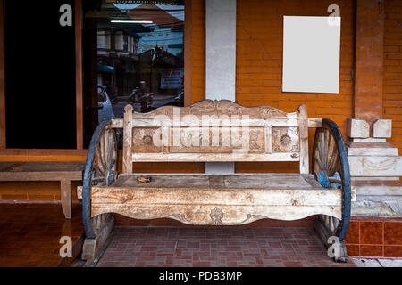 Vieux banc en bois trouvés à Bali avec design en bois et la roue de chariot les jambes. Contre un bâtiment en briques rouges avec une affiche et compte windows. Banque D'Images