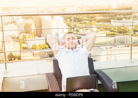 Satisfaits de travailler. Cheerful young man holding hands behind head et de garder les yeux fermés, assis sur la terrasse avec vue sur le paysage urbain Ba Banque D'Images