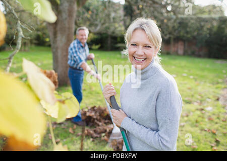 Portrait souriant, confiant mature woman raking autumn leaves in backyard Banque D'Images