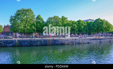 Shadwell Basin, Londres, UK - 7 mai 2018 : banque d'arbres avec un bon nombre de personnes bronzer et discuter. ligne de cycles le long du bord de l'eau. Banque D'Images