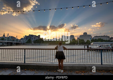 Londres, UK - 6 août 2018 : Une jeune femme sur la rive sud de la Tamise s'intéresse aux rayons crépusculaires comme le soleil se couche derrière Charing Cross Banque D'Images