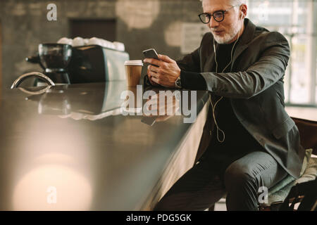 Mature businessman sitting at a cafe en utilisant le téléphone mobile. Man wearing earphones en regardant son portable tout en vous relaxant dans un coffee shop. Banque D'Images