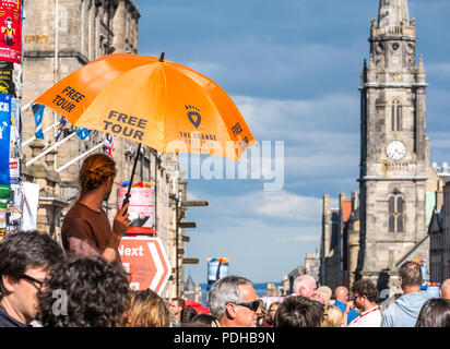 Edinburgh, Écosse, Royaume-Uni. 9 août 2018. Edinburgh Flinge Festival, Royal Mile, Édimbourg, Écosse, Royaume-Uni. Lors d'une journée de fête ensoleillée, le festival de rue est rempli de gens et d'artistes de la frange. Un homme tenant un grand parapluie orange annonçant la visite libre de moring se dresse au-dessus de la foule de piétons avec le tron Kirk spire en arrière-plan Banque D'Images
