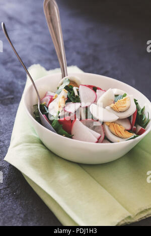 Vegan légumes salade de ramson, radis, les oignons verts et les oeufs durs avec yogourt dans une assiette blanche avec couverts sur une serviette sur une sombre concrète Banque D'Images