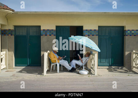 Un couple mature lire en dehors de leur cabine de plage sous un parasol sur la promenade des Planches de Deauville, Normandie, France Banque D'Images