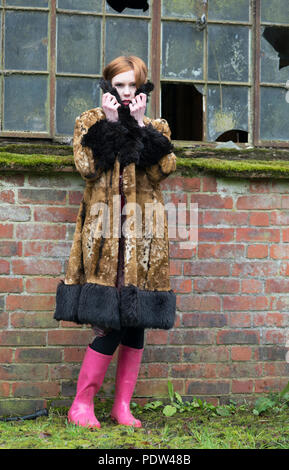 Red Headed girl in brown vintage fausse fourrure manteau avec garniture noire et bottes rose a l'air très froid en face d'un bâtiment de ferme à l'abandon de verre brisé. Banque D'Images