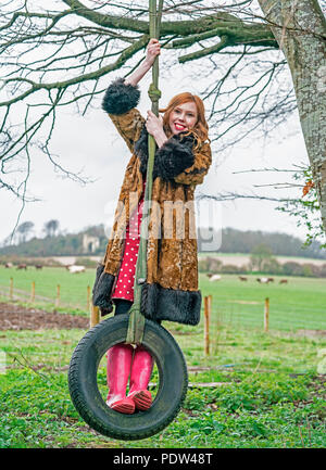 Red Headed dame en manteau de fourrure de faux vintage, robe rouge à pois rose et bottes wellington balançoires à partir d'un arbre sur un pneu en caoutchouc à la ferme. Banque D'Images