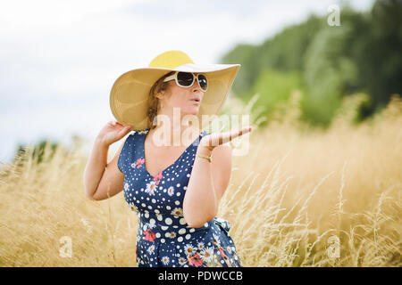 Femme d'âge moyen vêtus de blanc lunettes de soleil avec un chapeau à large chapeau, debout dans l'herbe longue whispy golden tout en soufflant un baiser par un beau jour Banque D'Images