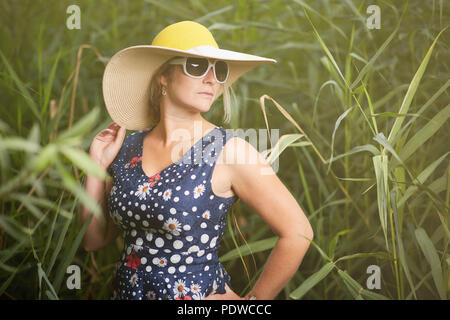 Femme d'âge moyen vêtus de blanc lunettes de soleil avec un large chapeau chapeau de l'article, dans l'herbe verte longue entre, regardant à droite Banque D'Images