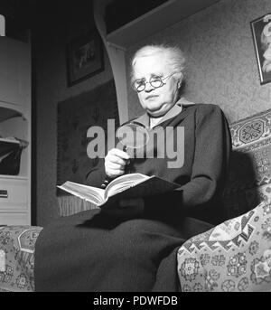 1940 femme avec des lunettes. Une vieille dame est à la recherche d'un livre en utilisant les deux verres et une loupe. La plupart des gens associent la loupe avec le caractère détective privé Sherlock Holmes créé par Conan Doyle. Il est également un symbole commun et de l'icône de clarifier les fonctions de recherche et de zoom dans les programmes d'ordinateur et sur internet. 1942. Kristoffersson Photo/A84-6 Banque D'Images