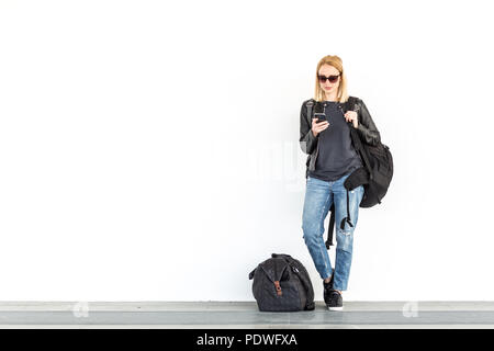 Woman en utilisant son téléphone portable en étant debout et en attente contre un mur blanc sur la station avec un sac de voyage à ses côtés. Banque D'Images