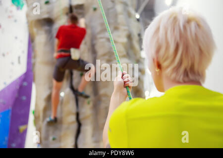Photo de retour de l'homme grimper sur un mur d'escalade et female trainer holding corde de sécurité dans la salle de sport Banque D'Images