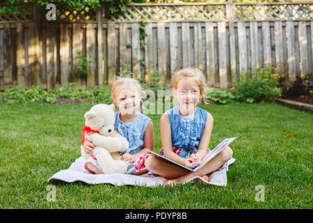 Portrait de groupe de deux enfants caucasiens blancs mignons amis assis sur l'herbe à l'extérieur avec réserve. Les filles d'âge préscolaire soeurs lecture livre sur l'arrière-cour. B Banque D'Images