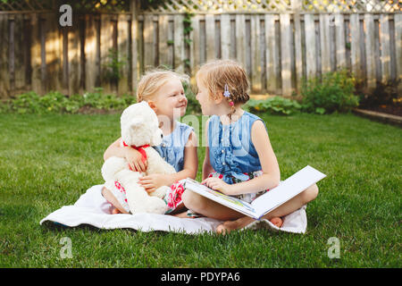 Portrait de groupe de deux enfants caucasiens blancs mignons amis assis sur l'herbe à l'extérieur avec réserve. Les filles d'âge préscolaire soeurs lecture livre sur l'arrière-cour. B Banque D'Images