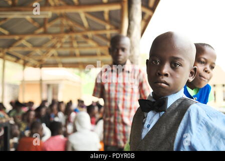 Le jeune garçon ougandais se tient dans son meilleur dimanche, en regardant à la caméra avec un groupe d'autres jeunes enfants africains qui attendent l'attention de l'hôpital Banque D'Images