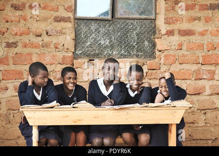 Les jeunes écoliers ougandais habillés dans un uniforme scolaire s'asseoir à l'extérieur sur des bancs en bois qui étudient pour leurs examens en classe avec de la brique en arrière-plan Banque D'Images