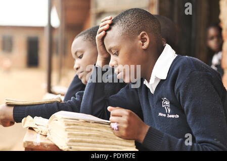 Les jeunes écoliers ougandais habillés dans un uniforme scolaire s'asseoir à l'extérieur sur des bancs en bois qui étudient pour leurs examens en classe avec de la brique en arrière-plan Banque D'Images