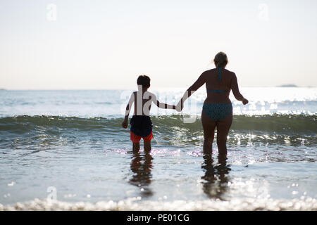 Mère et fils s'amusant de sauter les vagues de la mer en début de soirée alors que le soleil se couche et est sur le scintillement de l'eau d'un bleu pur Banque D'Images