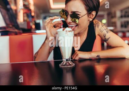 Femme portant des lunettes fantaisie assis dans un restaurant en compagnie de ses glaces milkshake. Femme souriante de lécher la garniture sur le milkshake à la th Banque D'Images