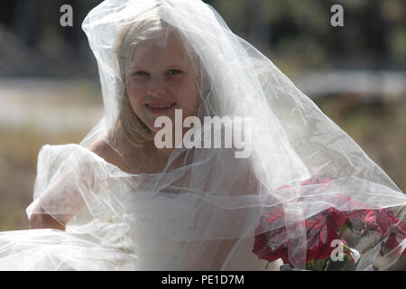 Fantasy, 8-9 ans blonde d'endroit, le port de la robe de mariée tante & voile. Assis en plein air sur grand rocher à côté de vase de roses rouges. Banque D'Images