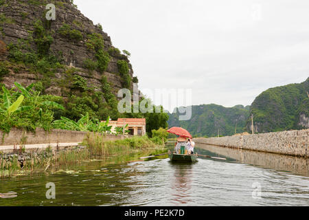 Un petit bateau en bois avec 3 passagers navigue dans un lac à Ninh Binh au Vietnam du Nord, avec les montagnes couvertes d'arbres en arrière-plan. Banque D'Images