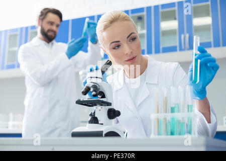 L'analyse en laboratoire. Belle jeune femme agréable assis devant le microscope et tenant un tube à essai en faisant une analyse de laboratoire Banque D'Images