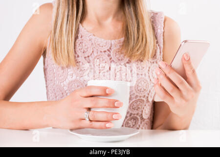 Belle jeune femme avec une tasse de café dans la main et smart mobile phone avec dentelle robe couleur crème. Mode et beauté concept. Close up Banque D'Images