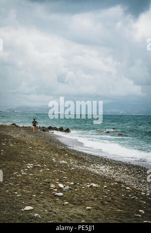 Scène de plage typique de l'île de Crète. De jeune femme marchant le long de la plage. Vue vers l'océan. Avec les montagnes en arrière-plan sur un ciel nuageux da Banque D'Images