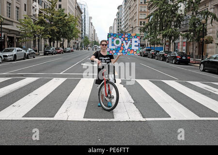 Washington D.C., États-Unis.12th août 2018.Une femme arrête son vélo sur un passage en croix sur 14th Street NW à Washington D.C., en tenant un panneau d'amour dans le cadre des manifestations de contre-manifestation contre le rassemblement « Unite the Right 2 » à Lafayette Park tout proche.Alors que les contre-manifestants se sont rendus dans la rue, au lieu d'avoir des barrières en place pour que les manifestants tournent à gauche au carrefour, cette femme seule s'y est levée pour soutenir les contre-manifestants en faisant un tour directement en face d'elle. Banque D'Images