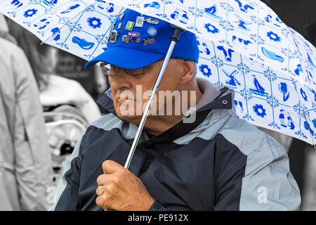 Senior man wearing cap avec insignes voiture s'abritant sous le parapluie dans la pluie à Bristol en Août Banque D'Images