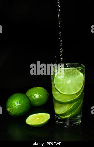 Les boissons rafraîchissantes d'été et concept. Jet de l'eau est versée dans le verre de boisson froide avec de la glace et tranche frais mûrs limes vert sur noir background Banque D'Images