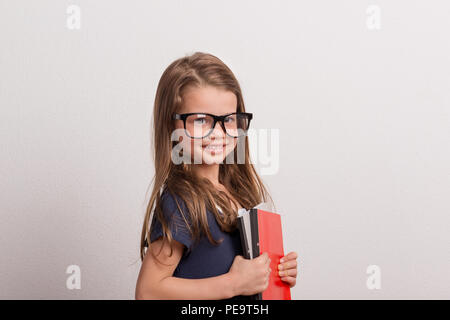 Portrait d'une petite écolière avec grands verres dans un studio, holding notebooks. Banque D'Images