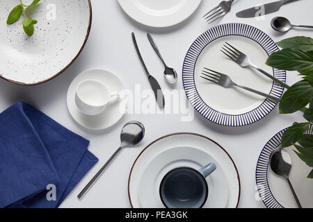 Vue de dessus de feuilles vertes, différentes plaques céramiques, tasse, essuie-tout, fourchettes, cuillères et couteaux de table blanc Banque D'Images