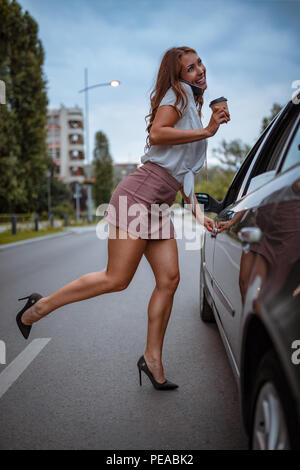 Une belle jeune femme dans une jupe courte parle sur le smartphone, tenant une tasse de café et s'apprête à entrer dans la voiture. Banque D'Images