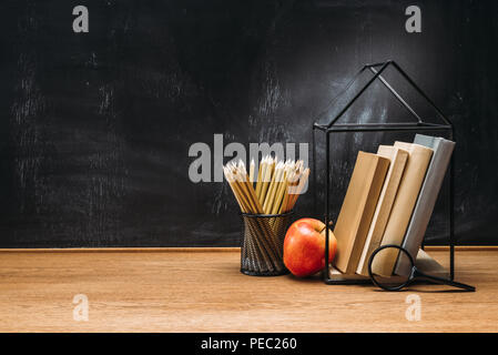 Vue rapprochée de pomme fraîche, loupe, des crayons et des livres sur la surface en bois avec tableau noir vide derrière Banque D'Images