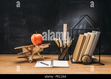 Vue en gros d'apple sur avion jouet en bois, ordinateur portable, des livres et des crayons sur la table d'examen avec tableau noir vide derrière Banque D'Images