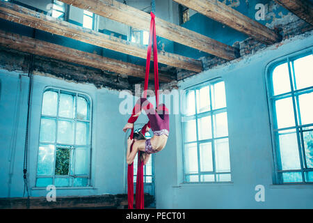 Gymnast gracieux exercice aérien avec des étoffes rouges sur fond bleu ancien loft. Young teen caucasian fit girl. Le cirque, acrobatie, Acrobat, interprète, sport, fitness, gymnastique concept Banque D'Images