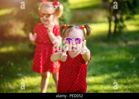 Deux mignonnes petites amies s'amuser dans la cour. petite fille afficher la vue bravo et bonne chance. vêtus de robes rouges lumineux et tendance sunglasse Banque D'Images