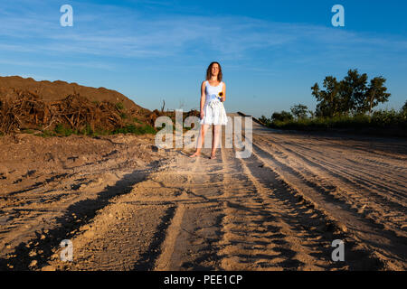 Déçu femme debout au milieu de la construction routière et de la tenue d'un tournesol cassé pendant le coucher du soleil. La destruction de l'environnement concept. Banque D'Images