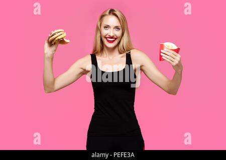 Belle jeune fille élancée sur un fond rose est titulaire d'un hamburger et des frites dans ses mains et sourit. Concept de la malbouffe Banque D'Images