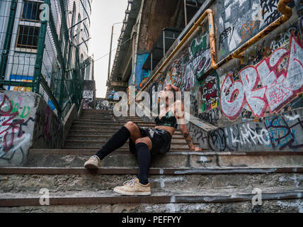 Plan large de street punk girl en milieu urbain. Fille assise sur l'escalier peint avec de l'écriture graffiti sous le pont Banque D'Images