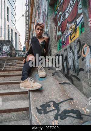 Plan large de street punk girl en milieu urbain. Fille assise sur l'escalier peint avec de l'écriture graffiti sous le pont Banque D'Images