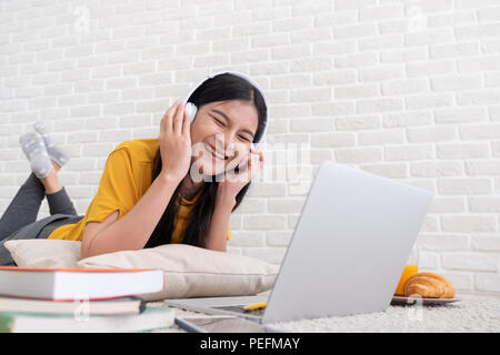 Femme d'origine asiatique d'écouter de la musique et utiliser un ordinateur portable pour travailler à la maison.femme se coucha sur tapis en mur de briques.en ligne de mode de travail Banque D'Images