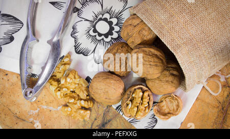 Photo horizontale de coques de noix et amandes dans sac de jute sur la surface en marbre avec serviette blanche avec des fleurs. Casse-noisette à côté de la noix. Banque D'Images