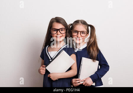 Deux petites écolières avec des lunettes et uniforme debout dans un studio, holding de blocs-notes Banque D'Images