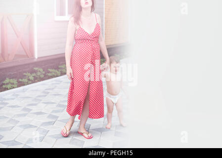 Jeune femme marche maman avec bébé fils marchant dans la rue joie happy smiling. Brunette en robe rouge style soleil fond flou copy space Banque D'Images