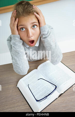 Petite lycéenne holding head avec bouche ouverte et fou de l'expression du visage. Photo de fille avec livre sur le bureau en classe. Retour à l'école Banque D'Images