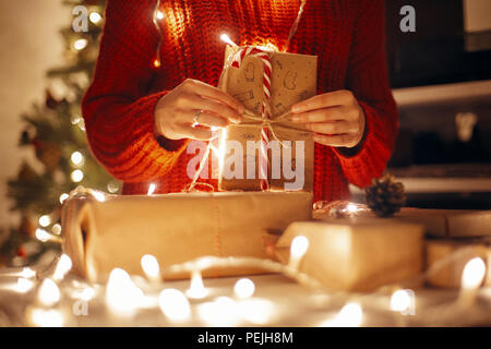 Moments magiques de noël. hands holding christmas present dans les lumières dans la salle des fêtes en soirée en vertu de l'arbre dans l'éclairage. espace pour texte. girl opening mo Banque D'Images