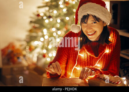Happy girl in santa hat et red sweater christmas lights en soirée dans la salle des fêtes à l'illumination de l'arbre d'ouverture des cadeaux d'artisanat moderne. Banque D'Images