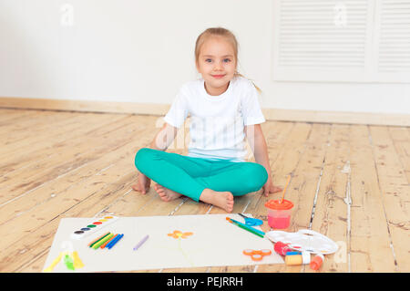 Petite fille blonde paintson big white paper assis sur le plancher à l'intérieur. Banque D'Images